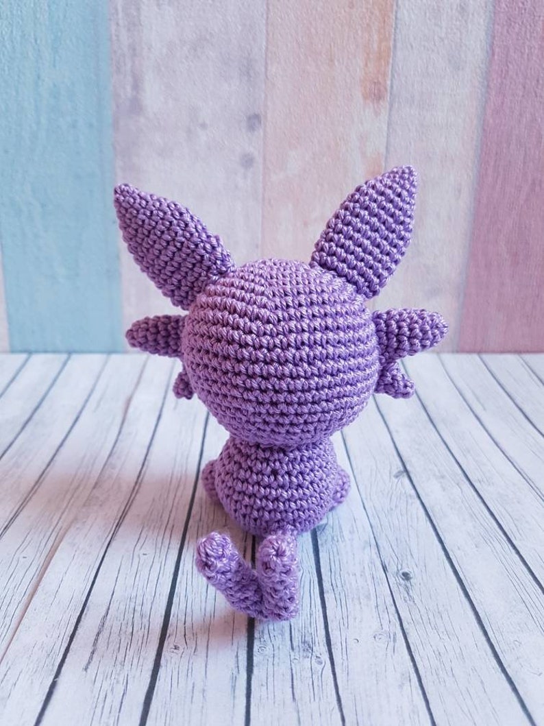 May include: A purple crocheted amigurumi toy in the shape of a cat sitting on a white wooden surface.