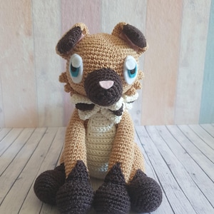 May include: A handmade crochet dog plushie in shades of brown, tan, and cream. The dog has dark brown paws, ears, and a snout, with light blue eyes and a pink nose. The plushie is sitting upright, showcasing its detailed crochet work.