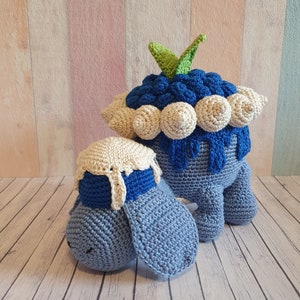 Puede incluir: Dos animales de peluche tejidos a crochet, uno azul con un gorro tejido a crochet blanco y el otro azul con un gorro tejido a crochet blanco y una hoja verde encima de un diseño de arándano tejido a crochet azul y blanco.