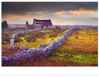 May include: A weathered stone wall snakes through a field of green and brown grasses, leading to a dilapidated stone building with a red roof. The sky is overcast with a hint of orange in the distance.