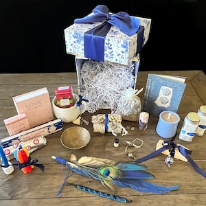 May include: A gift box with a blue floral pattern and a blue ribbon, filled with various items. Includes a candle, incense, a journal, and a peacock feather. The items are arranged on a wooden surface.