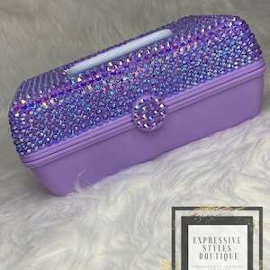 May include: A lavender-colored plastic jewelry box with a clear lid. The box is covered in iridescent rhinestones. The box has a white handle and a clear rhinestone button.