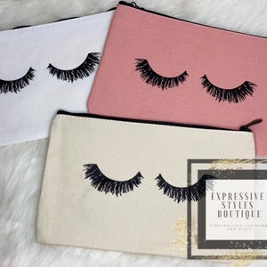 Könnte beinhalten: Drei Kosmetiktaschen aus Canvas mit schwarzen Wimpern bedruckt. Die Taschen sind weiß, beige und rosa. Die Taschen sind alle geschlossen und haben einen Reißverschluss.