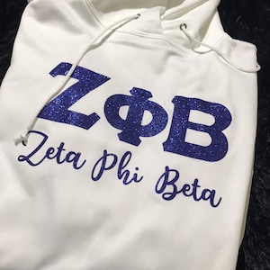 Puede incluir: Sudadera blanca con un diseño de purpurina azul que dice "ZΦB Zeta Phi Beta".