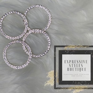 Puede incluir: Cuatro anillos plateados con diamantes de imitación transparentes. Los anillos están dispuestos en círculo sobre una superficie blanca y peluda. El texto "EXPRESSIVE STYLES BOUTIQUE PERSONALIZED CLOTHING AND GIFTS" es visible en el fondo.