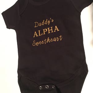 May include: Black baby onesie with gold lettering that reads "Daddy's ALPHA sweetheart".