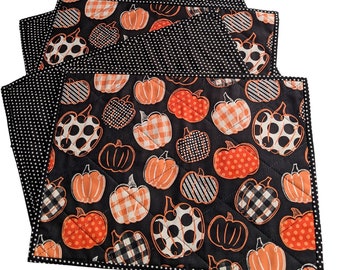 May include: Set of four quilted placemats. The placemats feature a black background with orange pumpkins in various patterns. The reverse side is black with white polka dots. The edges are trimmed with a black and white polka dot border.