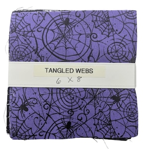 May include: A stack of square fabric pieces featuring a purple background with a black spiderweb and spider pattern. A white paper band wraps around the center, printed with the text TANGLED WEBS and the handwritten dimensions 6 x 8 inches.