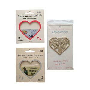 May include: Three packages of iron-on labels featuring heart-shaped designs. The first package is red and white with the text "Sweetheart Labels" and "Made with Love". The second package is brown and white with the text "Beatrix Potter Collection" and "Made with Mother's Love". The third package is white with the text "Christmas Trims" and a gold heart-shaped design.