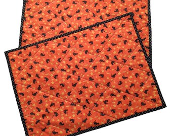 May include: Two orange and black quilted placemats with a repeating pattern of black cats and gold stars. The placemats have a black and white polka dot trim.