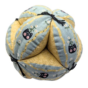 May include: A soft, spherical fabric toy constructed from alternating panels of light blue and yellow patterned cloth. The blue panels feature a repeating print of black cat faces with pink cheeks and white flowers, while the yellow panels have a subtle floral texture. Black thread secures the segments.