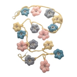 May include: A crocheted garland with pastel pink, blue, and cream flowers. The garland is made of yarn and is strung together with a cream-colored cord.