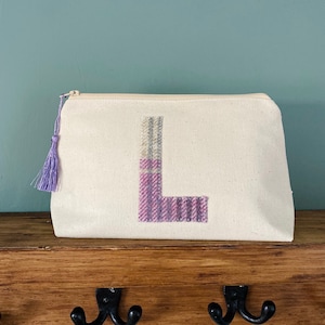 Op de afbeelding: Een witte canvas toilettas met een paarse en witte geruite letter L aan de voorkant. De tas heeft een paarse kwast.