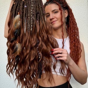 Puede incluir: Dreadlocks marrones y castaños con cuentas decorativas, plumas y detalles de cuero. Las rastas son sostenidas por una persona. Las rastas tienen una textura ondulada y son una mezcla de colores marrones y castaños.
