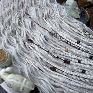 May include: White synthetic dreadlocks with brown and black beads and metal charms. The dreadlocks are arranged in a wavy pattern.