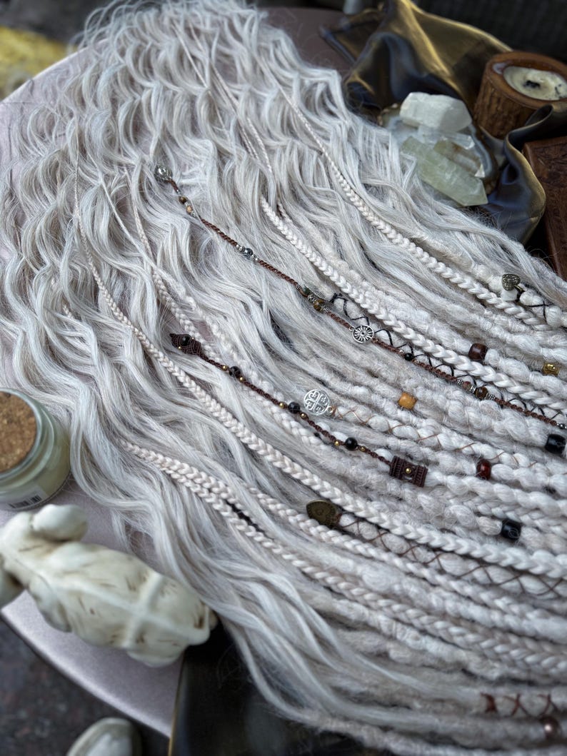 May include: White synthetic dreadlocks with brown and black beads and metal charms. The dreadlocks are arranged in a wavy pattern.