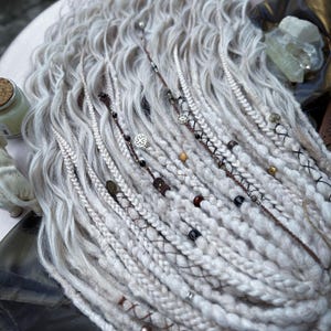 May include: White synthetic dreadlocks with brown, black, and silver beads and charms. The dreadlocks are long and wavy.