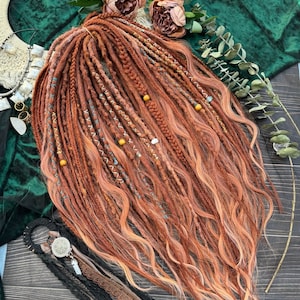 May include: Copper-coloured dreadlocks with light orange tips, decorated with blue and white thread accents and small beads. The dreadlocks are styled with braids and loose waves. The image also includes decorative accessories and flowers.
