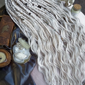 May include: White synthetic dreadlocks with beads and charms, displayed on a brown fabric surface with crystals and a small elephant figurine.