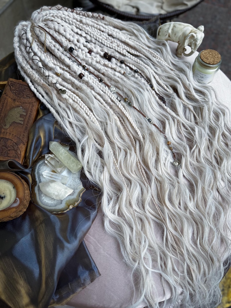 May include: White synthetic dreadlocks with beads and charms, displayed on a brown fabric surface with crystals and a small elephant figurine.
