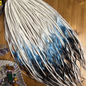 Puede incluir: Dreadlocks sintéticas ombre en blanco, azul y negro. Las dreadlocks son largas y onduladas.