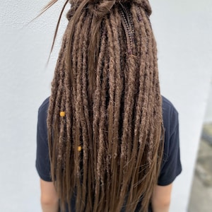 May include: Brown synthetic dreadlocks with small yellow beads. The dreadlocks are pulled back into a ponytail.
