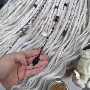 May include: White synthetic hair extensions with brown and black beads and charms. The hair is braided and tied with brown string. A small brown and black charm is held in a hand.