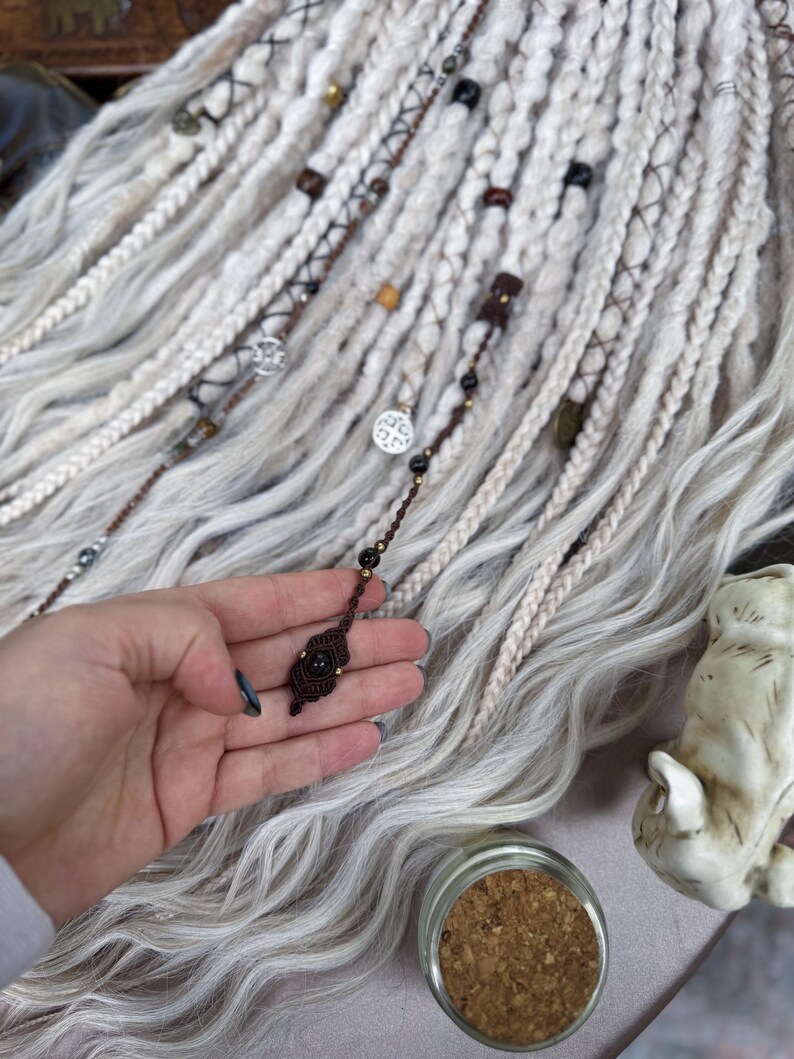May include: White synthetic hair extensions with brown and black beads and charms. The hair is braided and tied with brown string. A small brown and black charm is held in a hand.