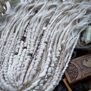 May include: White and light brown synthetic dreadlocks with beads and charms. The dreadlocks are arranged in a fan shape and are lying on a dark surface.