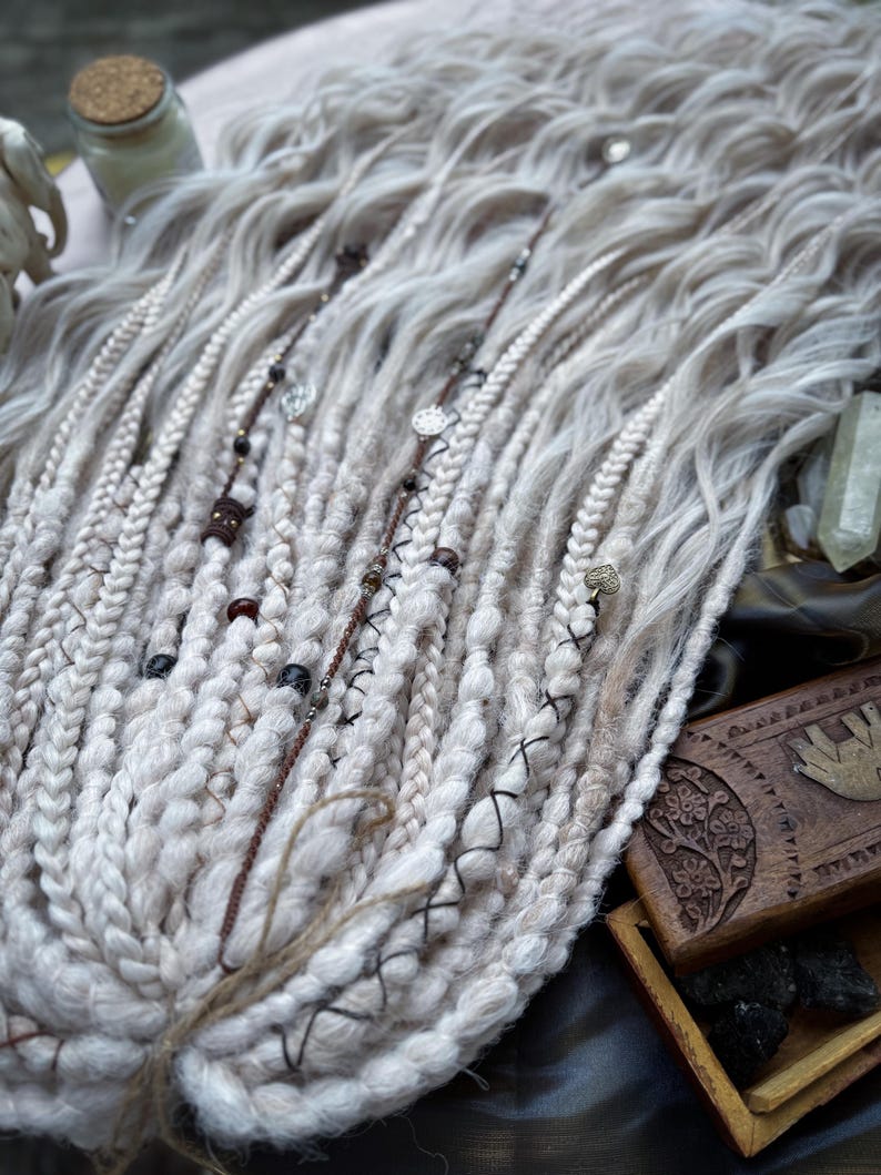 May include: White and light brown synthetic dreadlocks with beads and charms. The dreadlocks are arranged in a fan shape and are lying on a dark surface.