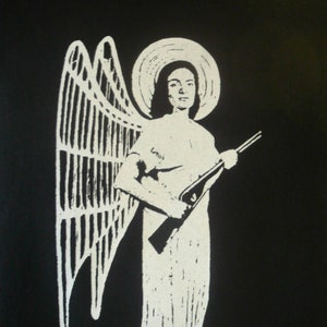 以下が含まれることがあります： 白い天使がライフルを持っている白いプリントが施された黒いTシャツ。天使は後光と翼を持っている。天使の胸には「Lord」という文字がプリントされている。