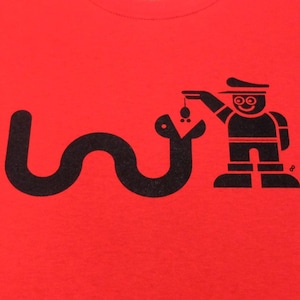 Pode incluir: Camiseta vermelha com um gráfico preto de uma cobra, uma pessoa com um chapéu e um pequeno objeto. A pessoa está segurando o objeto para a cobra. A camiseta é exibida em um manequim.