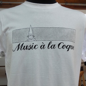 Könnte beinhalten: Weißes T-Shirt mit schwarzem Grafikdesign. Das Design zeigt eine Cartoonfigur mit einer Dummkappe in einem rechteckigen Rahmen, darunter der Schriftzug "Music à la Coque" in Kursivschrift. Das T-Shirt hat kurze Ärmel und einen Rundhalsausschnitt.