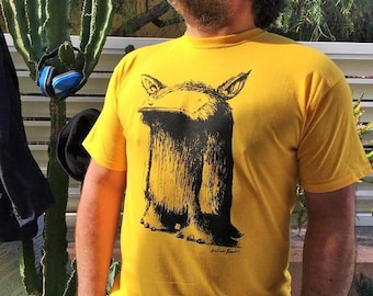 Könnte beinhalten: Ein gelbes T-Shirt mit einer schwarz-weißen Illustration eines Wesens mit einer langen Schnauze und großen Füßen. Das Wesen trägt einen Hut. Das T-Shirt trägt die Unterschrift des Künstlers unten rechts.