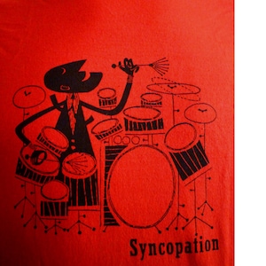 Op de afbeelding: Rood T-shirt met een zwarte illustratie van een cartoon-drummer die een drumstel bespeelt. De illustratie bevat een drumstel met bekkens, een basdrum en een snaredrum. De tekst "Syncopation" is onder de illustratie gedrukt.