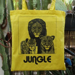 Peut inclure: Sac fourre-tout jaune avec une illustration noire d'un lion et de deux lionnes. Le mot "JUNGLE" est imprimé en noir sous l'image. Le sac a des poignées jaunes et est fait d'un matériau semblable à de la toile.