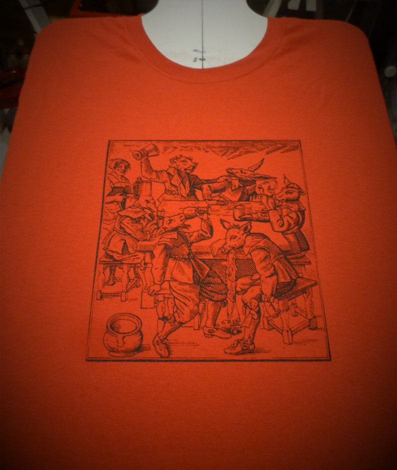 May include: A black and white illustration of a group of anthropomorphic animals drinking and socializing in a tavern setting. The animals are dressed in medieval clothing and are holding mugs of beer. The illustration is printed on a red t-shirt.