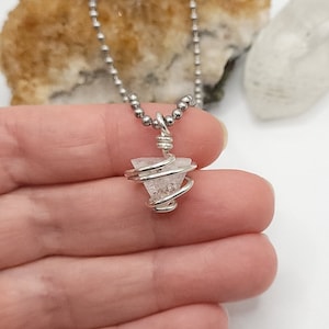 May include: A silver-toned necklace featuring a small, clear crystal pendant wrapped in wire. The crystal has a triangular shape and is suspended from a delicate ball chain. The pendant is the focal point, showcasing the natural beauty of the crystal.
