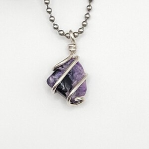 May include: A silver chain necklace with a purple and black charoite gemstone pendant wrapped in silver wire.