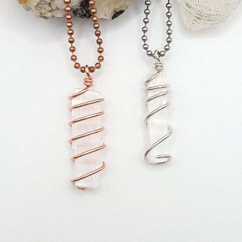 May include: Two necklaces with rectangular, clear crystal pendants. One pendant is wrapped in copper wire and hangs from a copper ball chain. The other pendant is wrapped in silver wire and hangs from a silver ball chain. The background is white.
