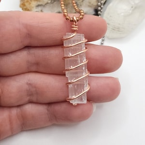 May include: A rectangular, light pink crystal pendant wrapped in copper wire, suspended from a beaded copper chain. The crystal is a pale, translucent color, and the wire is tightly wound around it. The necklace is displayed against a neutral background.