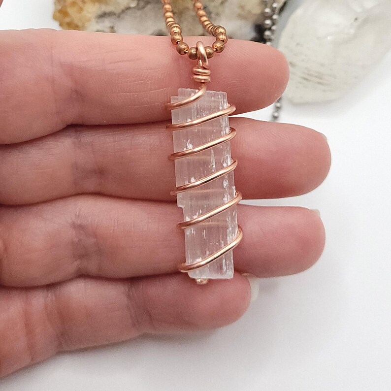 May include: A rectangular, light pink crystal pendant wrapped in copper wire, suspended from a beaded copper chain. The crystal is a pale, translucent color, and the wire is tightly wound around it. The necklace is displayed against a neutral background.
