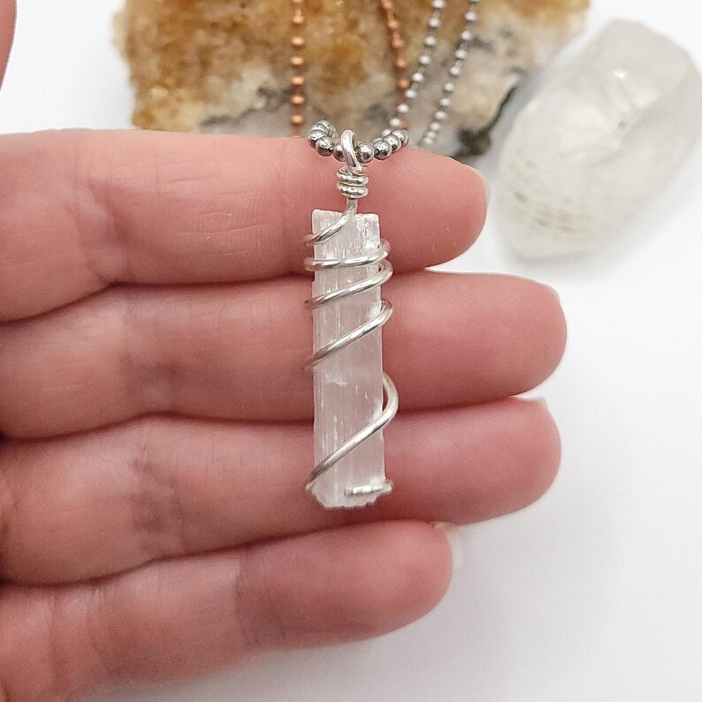 May include: A selenite crystal pendant wrapped in silver-colored wire, suspended from a beaded chain. The rectangular crystal is a pale, translucent white. The wire is coiled around the crystal. The background includes other crystals and a hand.