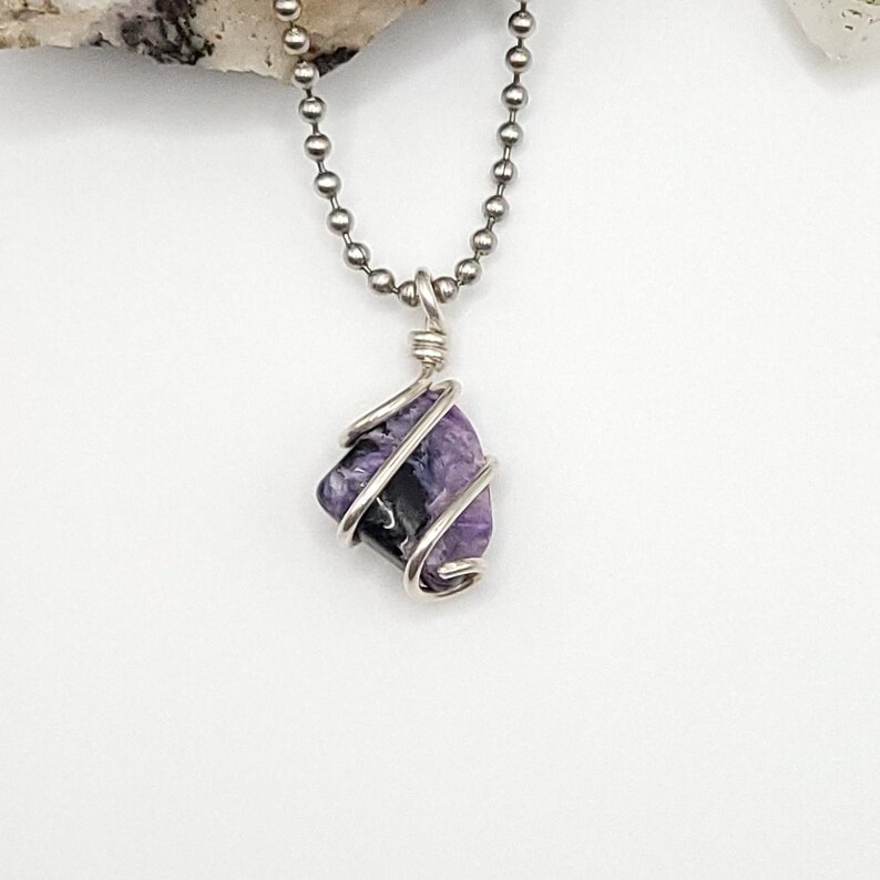 May include: A silver chain necklace with a purple and black Charoite gemstone pendant wrapped in silver wire.