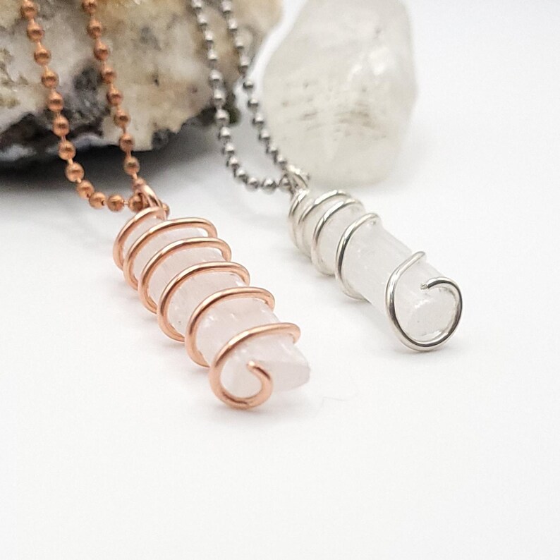 May include: Two crystal pendant necklaces. One features a pink crystal wrapped in copper wire on a copper chain. The other has a clear crystal wrapped in silver wire on a silver chain. Both crystals are cylindrical.