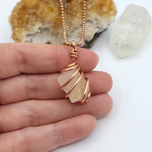 May include: A copper wire-wrapped pendant necklace featuring a raw, light yellow crystal. The pendant hangs from a delicate copper beaded chain. The crystal is encased in a spiral design.