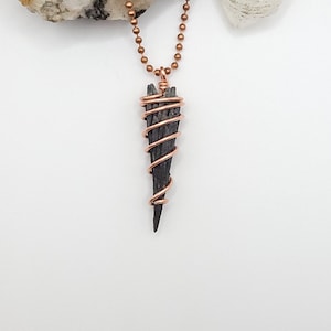 May include: A handmade pendant necklace featuring a dark, elongated stone wrapped in copper wire. The stone is suspended from a copper ball chain. The pendant has a triangular shape and is set against a white background.