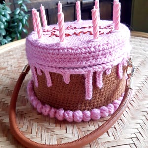 May include: A crocheted cake-shaped handbag with pink frosting and brown cake layers. The bag has a brown handle and is topped with pink candles. The word "Happy" is written on the top of the cake.