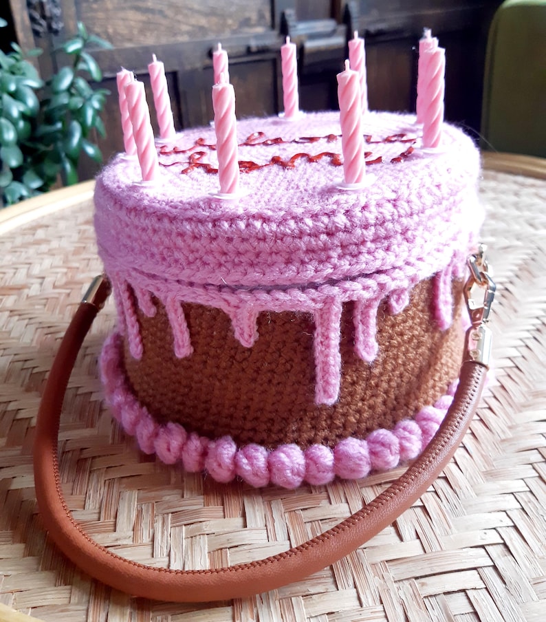 May include: A crocheted cake-shaped handbag with pink frosting and brown cake layers. The bag has a brown handle and is topped with pink candles. The word "Happy" is written on the top of the cake.