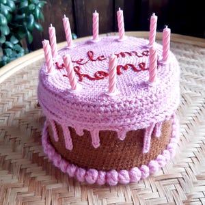 May include: A crocheted cake with a brown base and a pink top, adorned with pink candles. The top of the cake has the words "welcome home" embroidered in red. The cake sits on a woven tray.